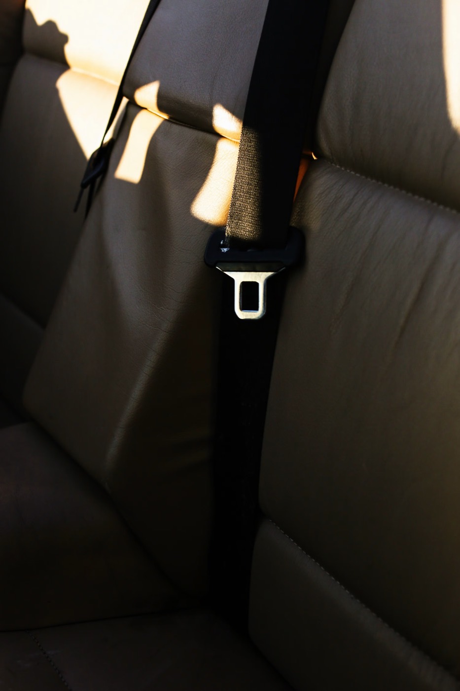 A seatbelt tongue plate and webbing against a tan leather rear seat, lit by low directional sunlight.