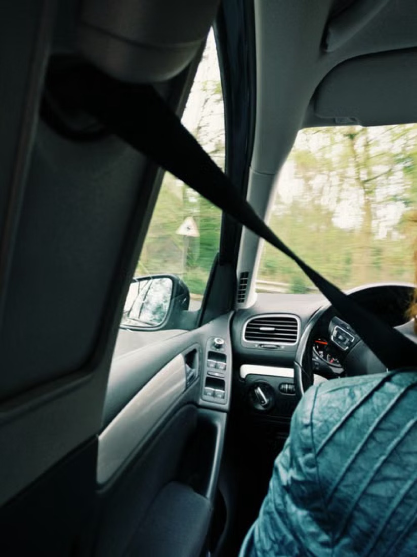 Interior view of a moving vehicle with the shoulder belt drawn across the occupant and open road visible through the windows.